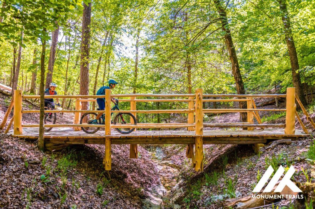 two people riding bikes on a wooden bridge in the woods.