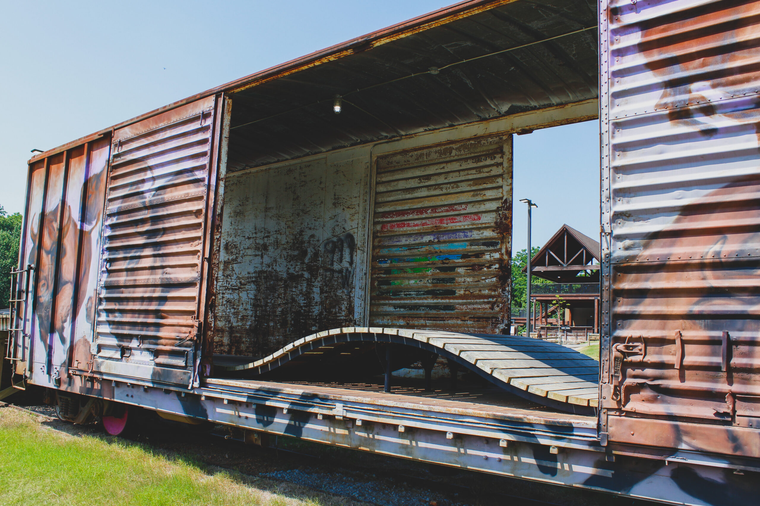 Graffiti-covered, rusted boxcar with open doors reveals a wavy wooden structure inside, set against a bright sky and distant structure.