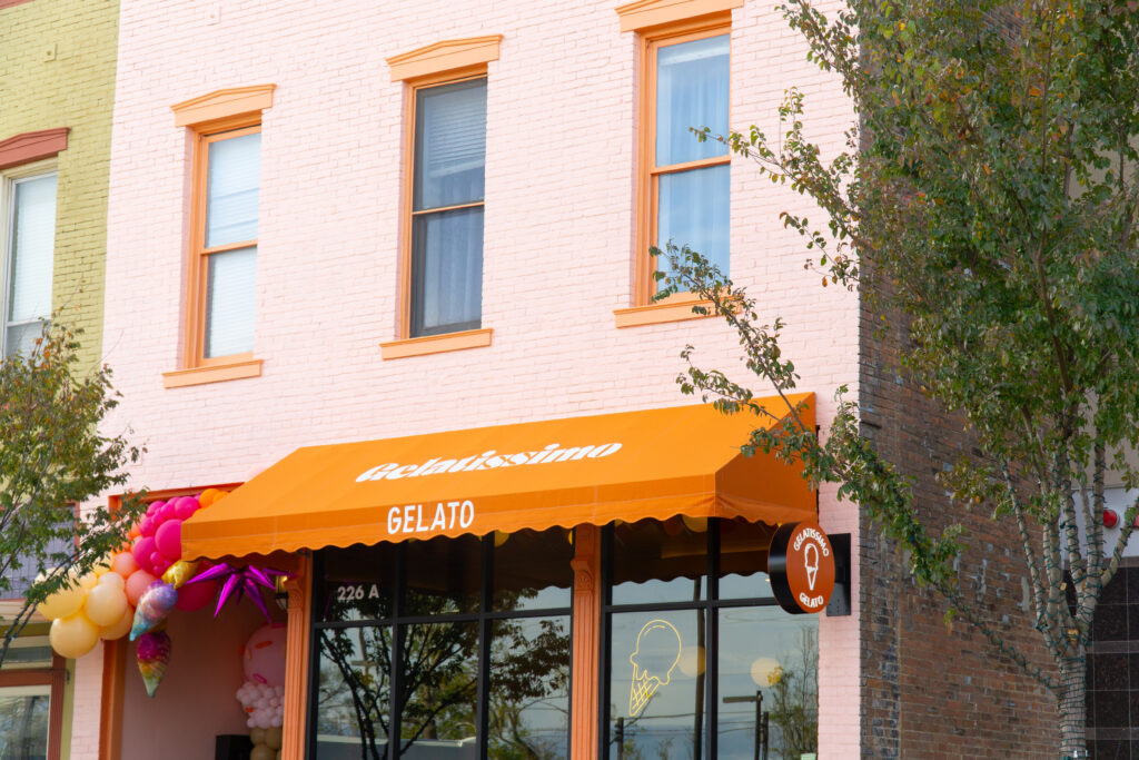 A pink brick building with an orange awning reads "Gelatissimo Gelato." Balloons are attached to the side of the awning. Tree branches are visible on the right.