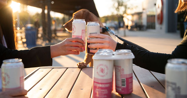two people toasting at a table with cans of beer.