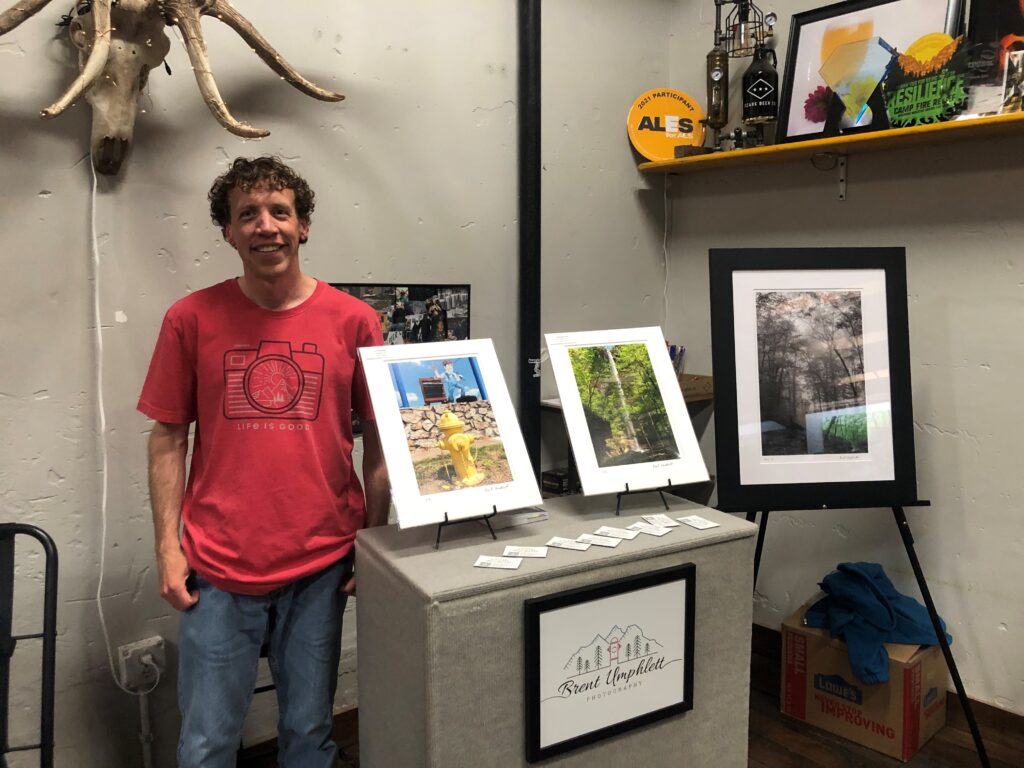 A person in a red shirt stands next to a display of framed photographs on easels. The display is atop a box with a logo. There are photos and decor items on the wall behind.