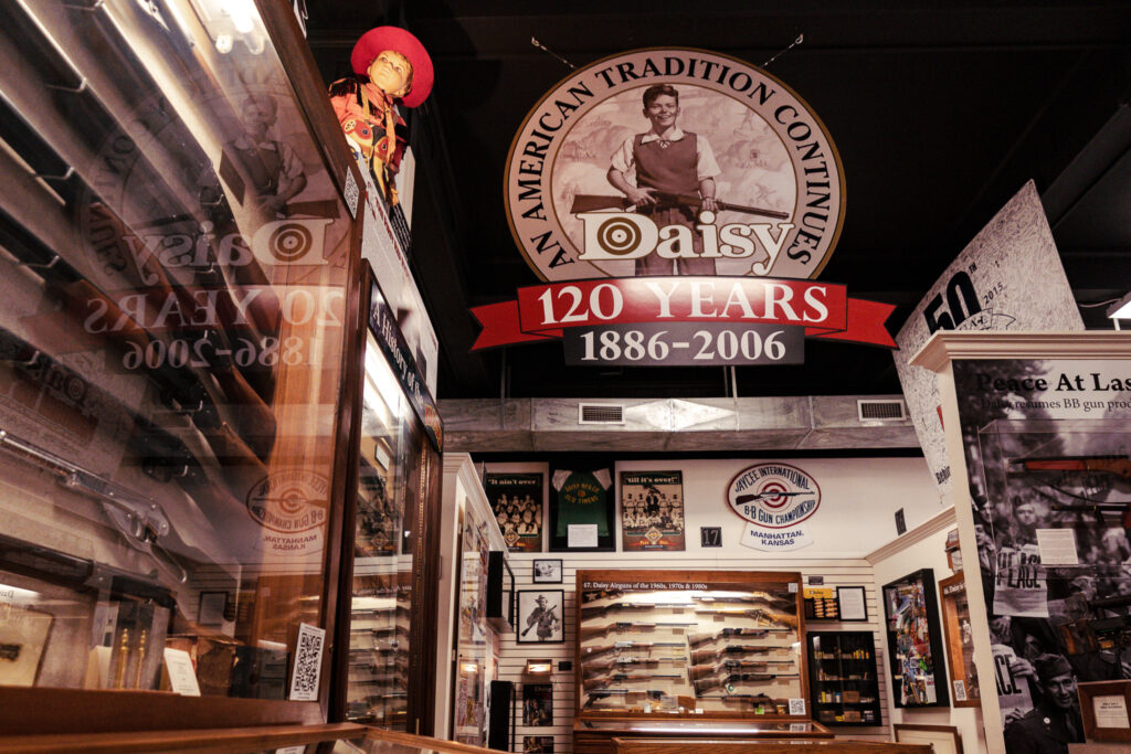 Museum exhibit featuring a large "Daisy 120 Years 1886–2006" sign, display cases with BB guns, and historic memorabilia celebrating the Daisy brand's history.