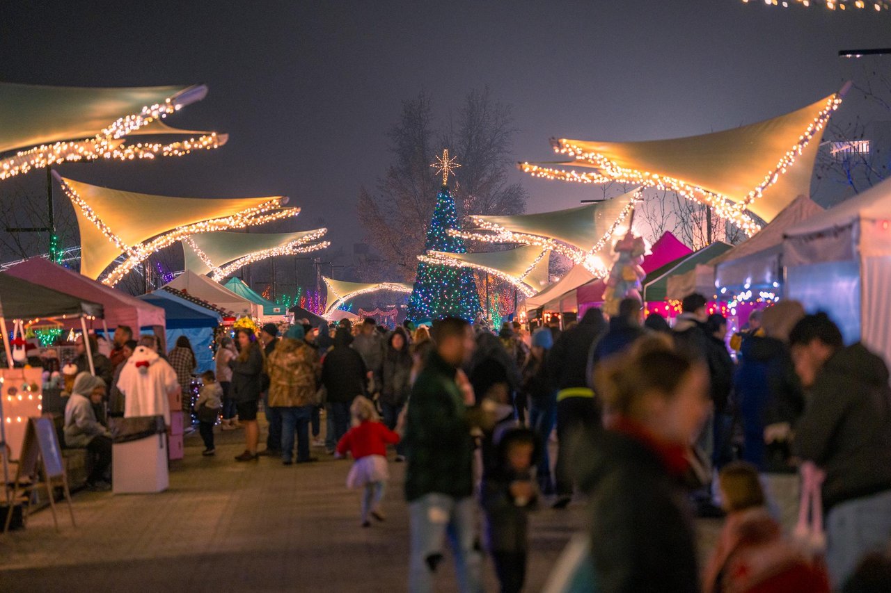 Crowd of people at an outdoor holiday market at night, decorated with string lights and a large Christmas tree.