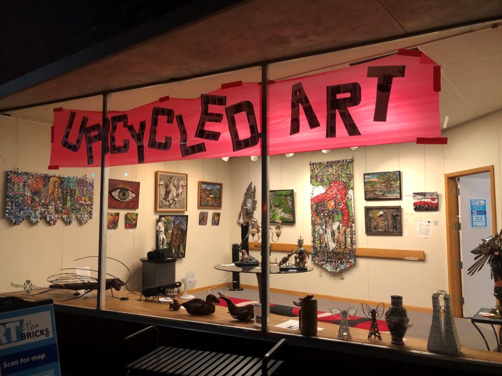 Storefront window displaying upcycled art pieces. A pink banner with "Upcycled Art" is above the window.