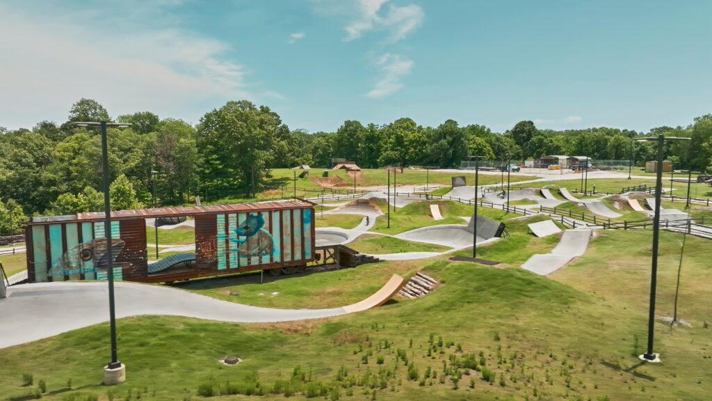 Outdoor skate park with winding concrete paths, grassy areas, and a mural on a shipping container. Surrounded by trees under a clear blue sky.