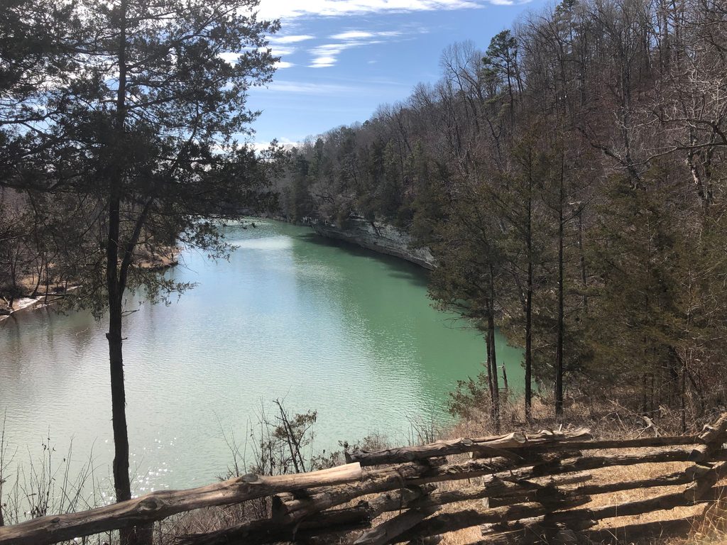 A green river winds through a wooded valley, viewed from behind a split-rail wooden fence.