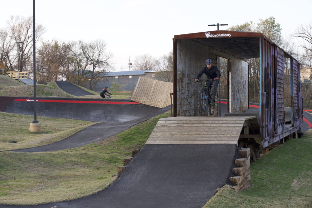 Cyclist rides through a wooden structure on a BMX track, while another cyclist is seen in the background.