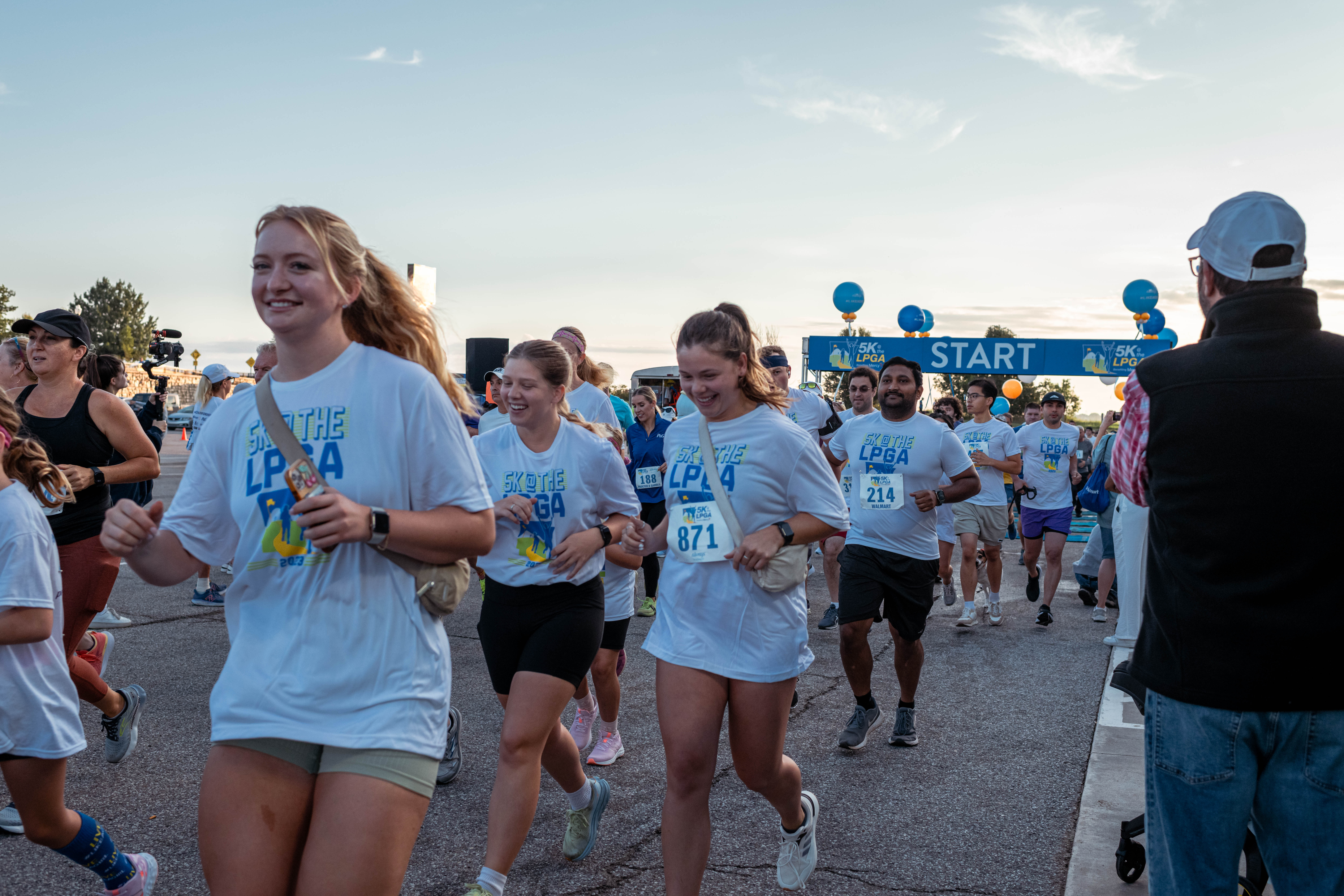 A group of people in athletic wear participate in a running event, passing under a starting line banner in an outdoor setting.