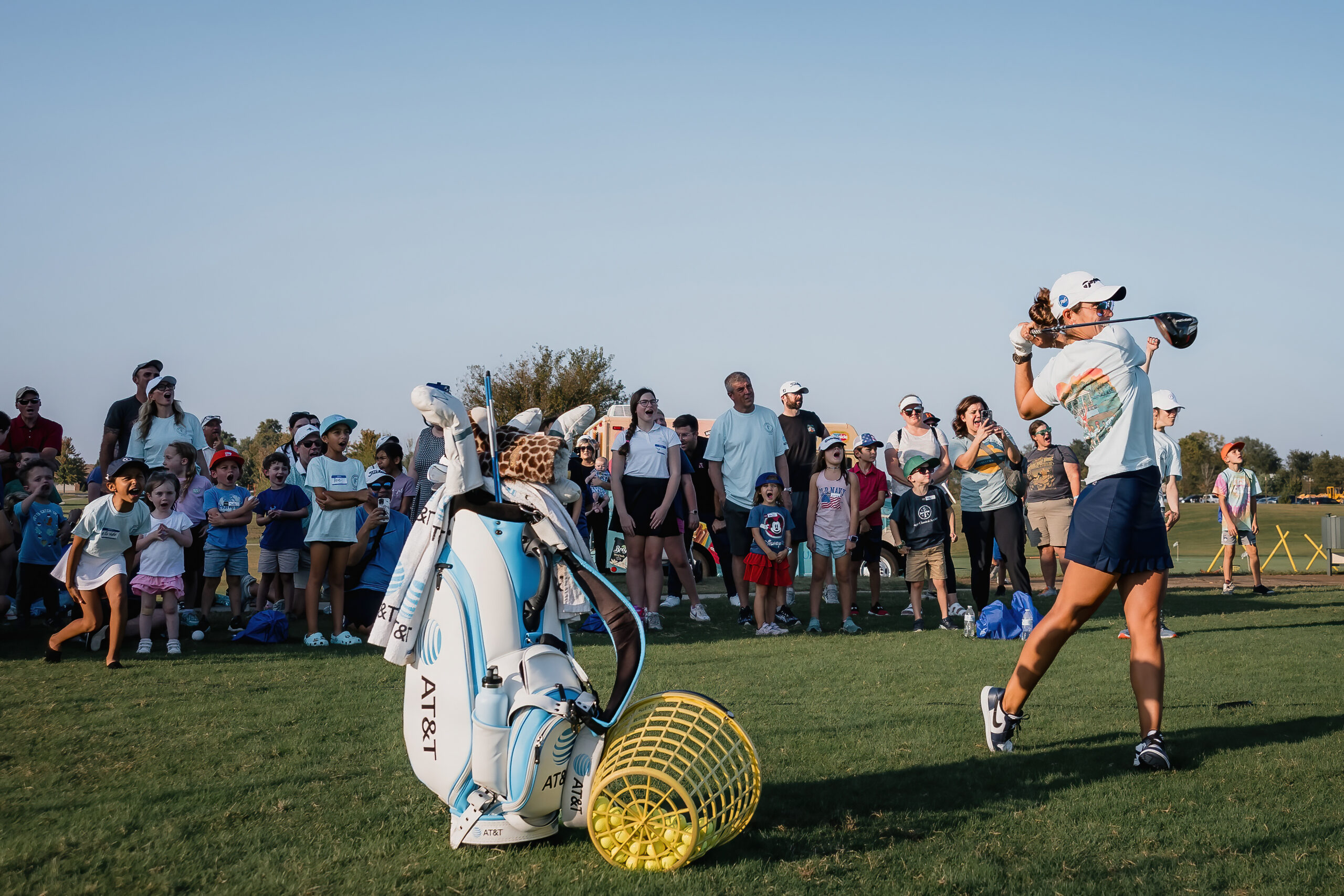 A golfer, viewed in profile, swings a club on a grassy field with a crowd of spectators in the background. A golf bag and a basket of balls are in the foreground.
