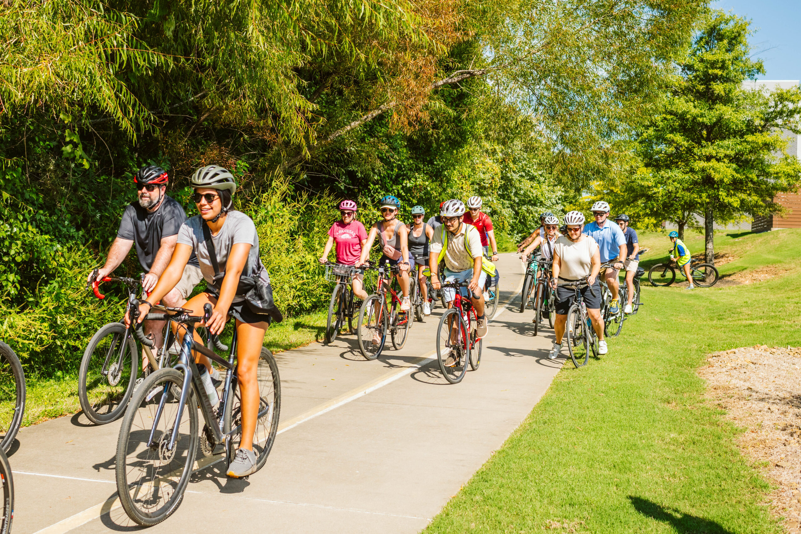 A group of cyclists wearing helmets rides along a paved path surrounded by greenery on a sunny day.