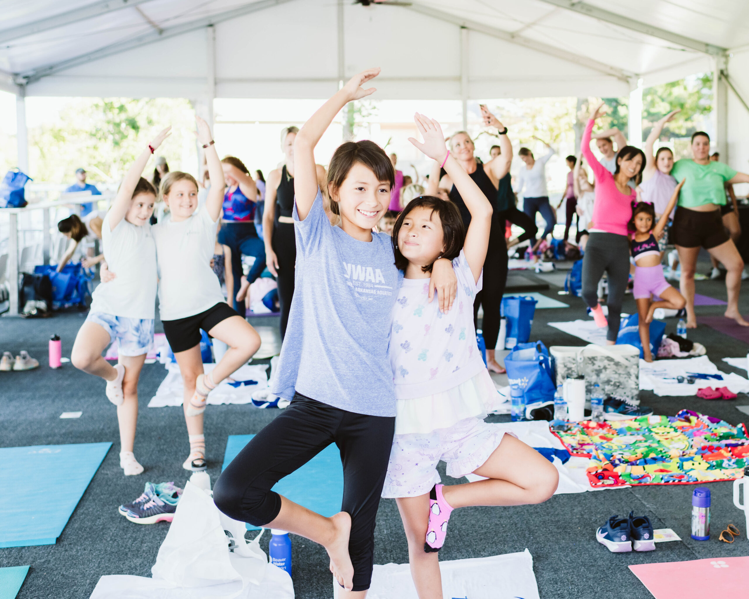 A group of people, including children, participate in a yoga class inside a large, white, tented structure. Many are balanced on one leg with arms raised. Yoga mats and water bottles are visible on the ground.