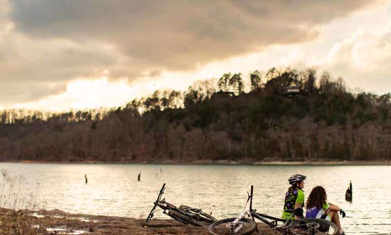 Two cyclists rest by Beaver lakeshore with their bikes lying on the ground. Forested hills are in the background under a cloudy sky.