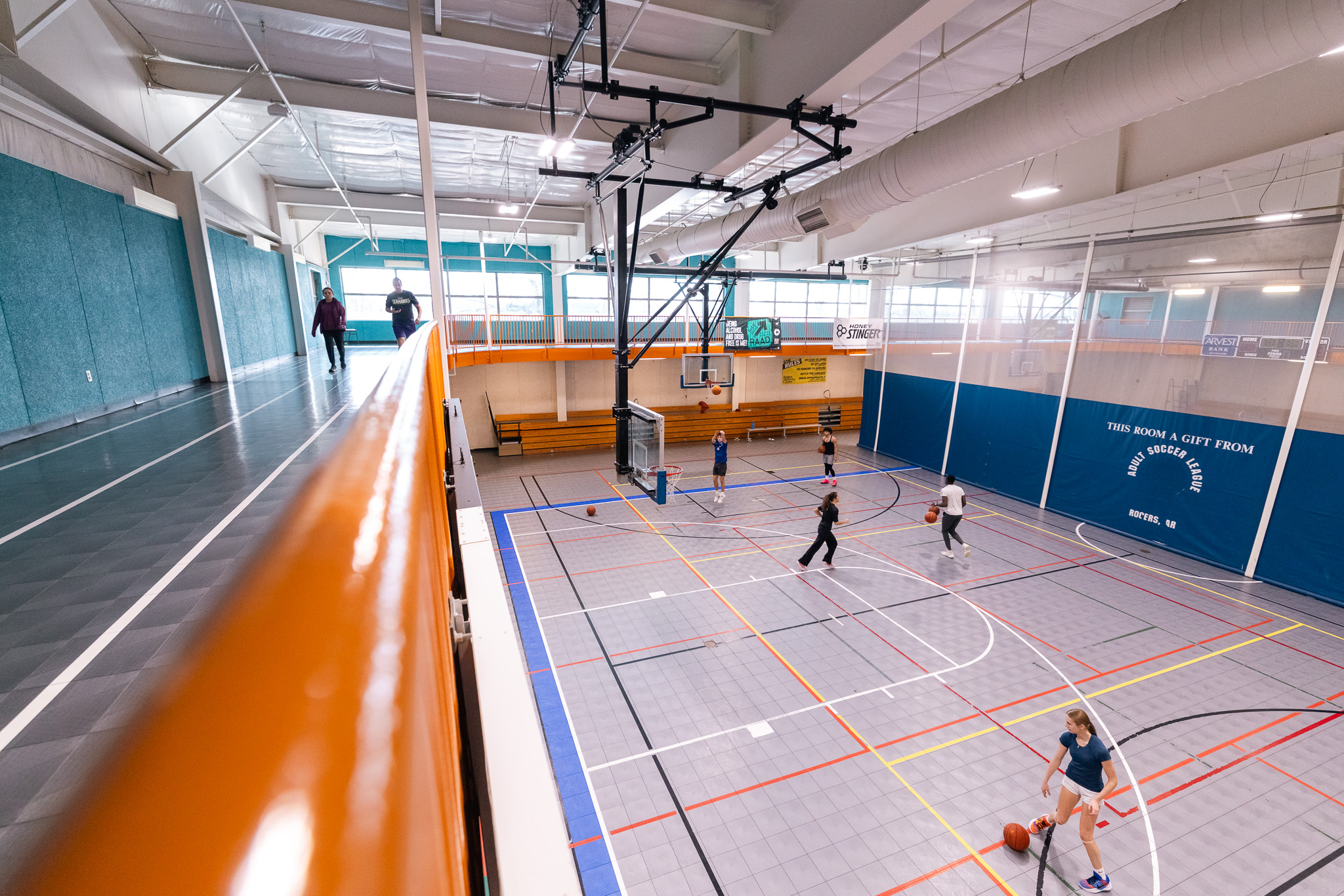 People play basketball on an indoor court while others walk along a second-floor track overlooking the gym. The gym has bright lighting, bleachers, and windows.