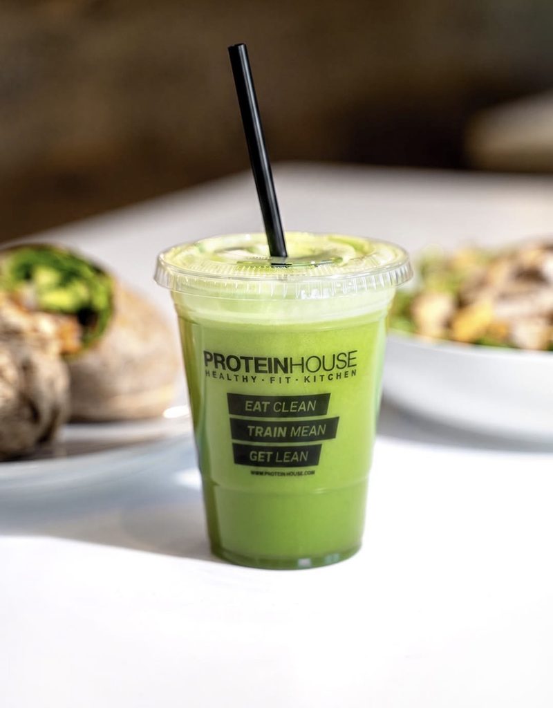 A plastic cup of green juice with a black straw sits on a table, labeled “Proteinhouse” with the phrases “Eat Clean, Train Mean, Get Lean.” Food dishes are blurred in the background.