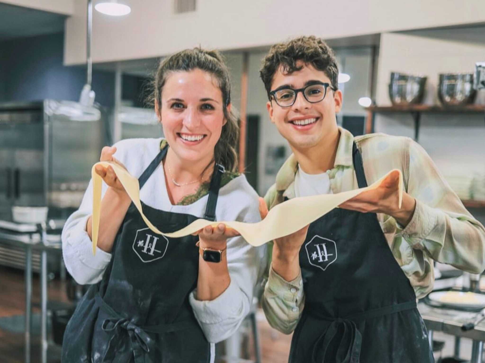 Two people in aprons hold a strip of pasta dough in a kitchen.