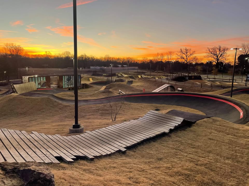 A bike park with curved wooden paths and ramps under a colorful sunset sky. Lamp posts and trees are visible in the background.