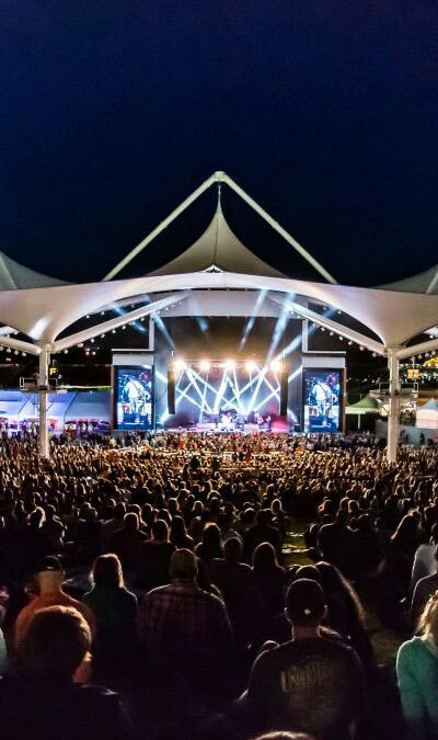 Large crowd attending an outdoor concert at night under a canopy, with bright stage lights and screens displaying the performers.