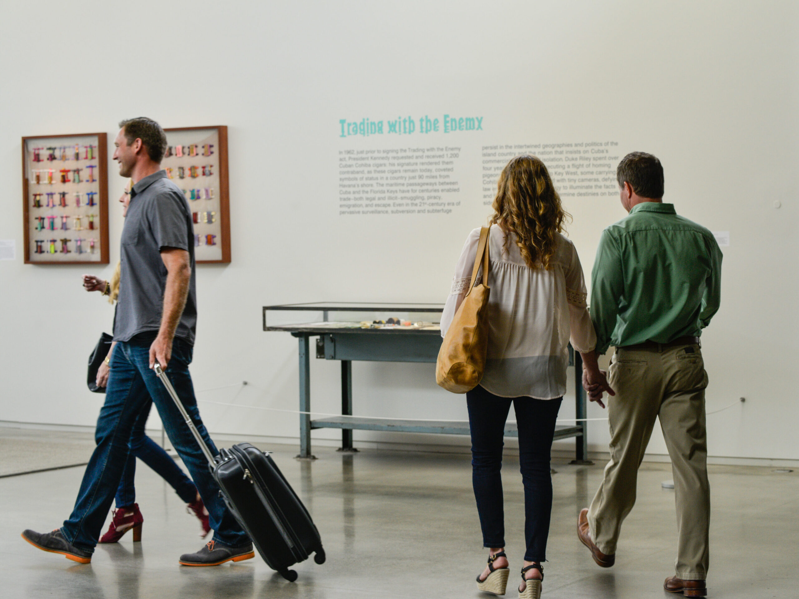 Three people walking in an art gallery. One person pulls a suitcase, while the other two hold hands. Artwork and text are displayed on the walls.