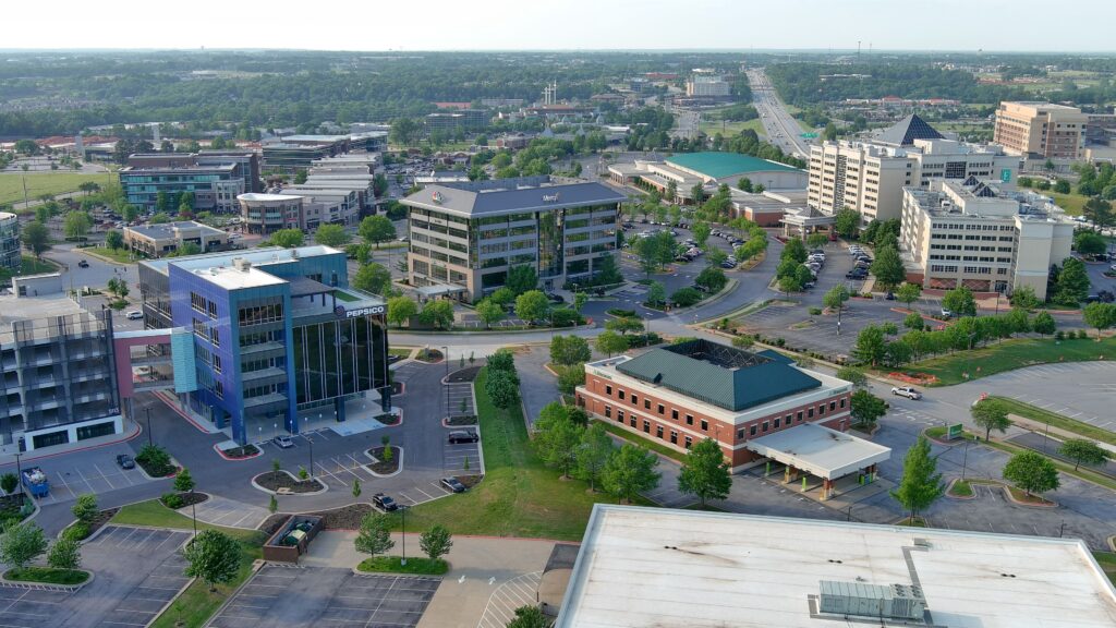 Aerial view of a convention district with multiple office buildings, roads, and parking lots, surrounded by green trees and grass under a clear sky.