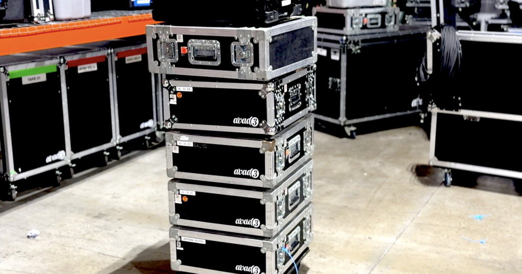 A stack of flight cases with metal edges and latches, labeled with "atoms," standing on a warehouse floor surrounded by other equipment cases.