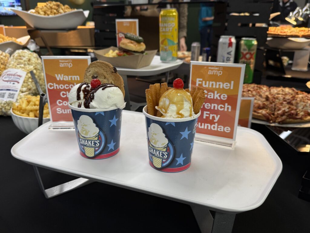 Two cups of sundaes; one with chocolate syrup, cookies, and a cherry, and the other with caramel, funnel cake fries, and a cherry. Various snacks and canned drinks are in the background.