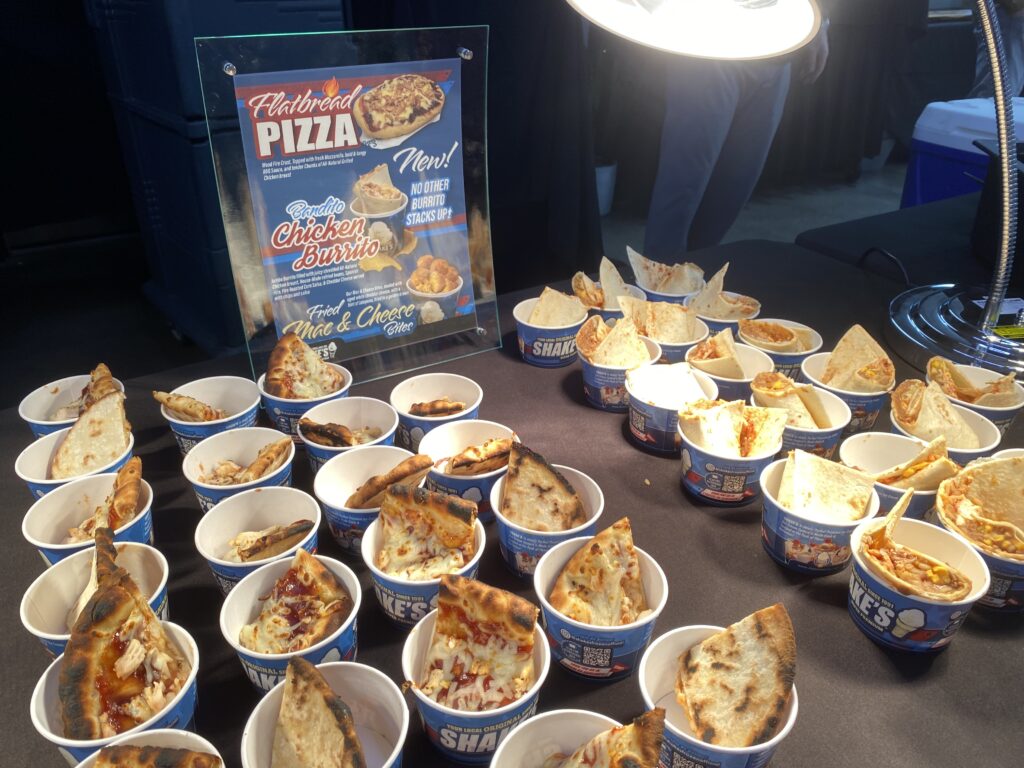 Cups of flatbread pizza samples on a table next to a promotional sign under bright lighting.