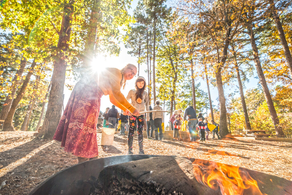 Two people roast food over a fire pit in a sunlit forest, with a group of others gathering in the background among tall trees.