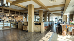 A spacious, modern restaurant interior with wooden beams, open kitchen, large windows, and booths along one side. A bar area is on the right, with a few people present.