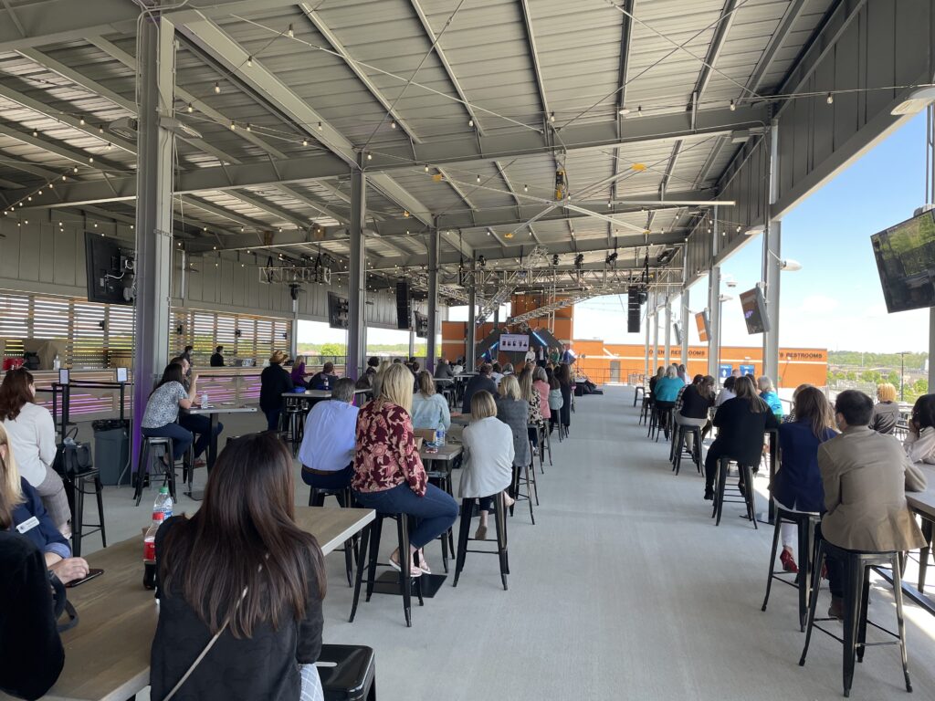 People seated on benches and stools under a covered outdoor pavilion, watching a presentation or event at the front.