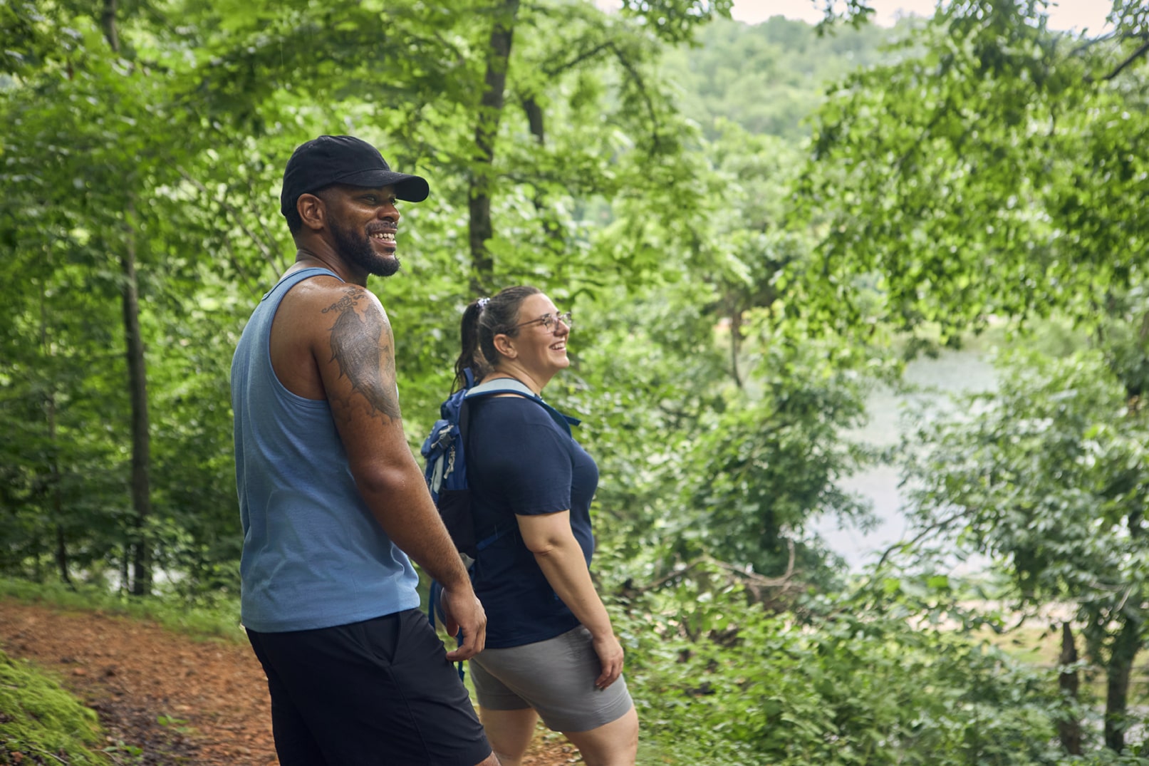 Two people, one man and one woman, hike on a forest trail surrounded by green trees and foliage, smiling and looking at the scenery.