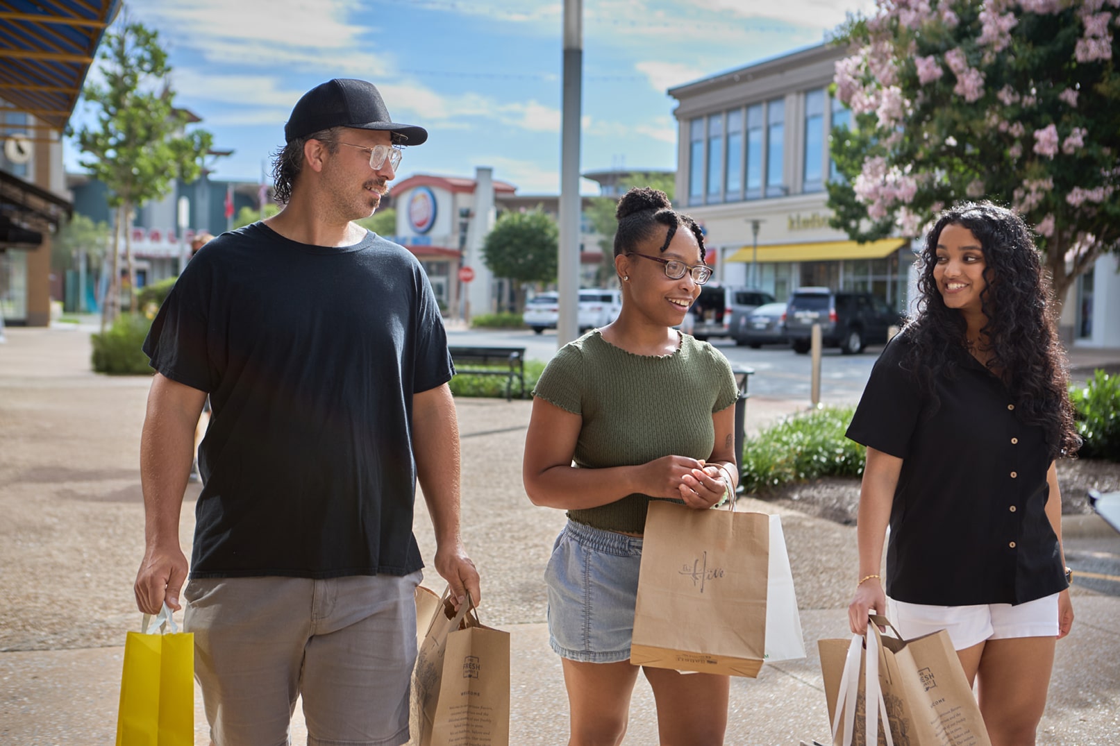 Three people walk outdoors in a shopping area, each holding paper shopping bags and engaging in conversation on a sunny day.