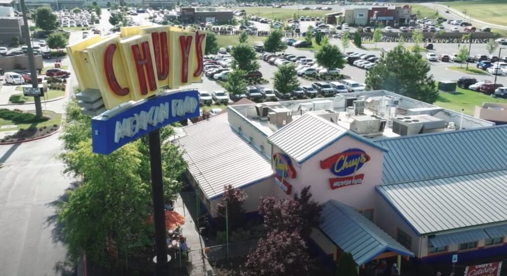 Aerial view of a Chuy's Tex-Mex restaurant with a large sign, parking lot, and nearby buildings in the background.