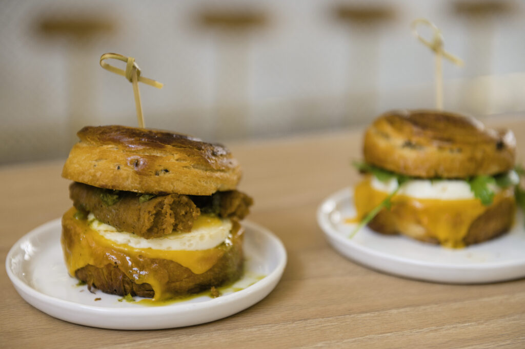 Two breakfast sandwiches with melted cheese, fried patty, egg, and greens on brioche buns, each secured with a wooden pick, served on small white plates.