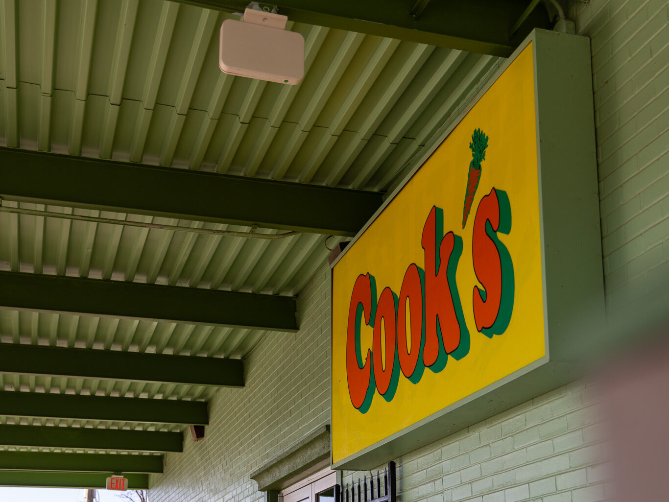 A yellow sign with "Cook's" in red and green letters hangs above the entrance of a light green brick building with metal railings.
