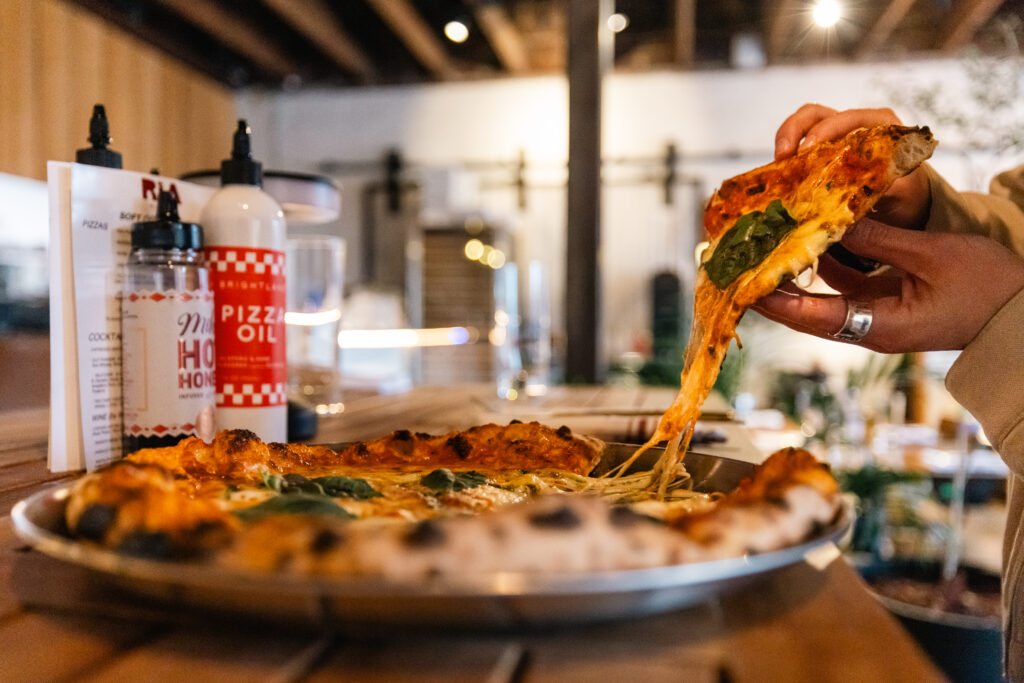 Slice of Margherita Pizza being pulled from a full pie inside Ria Pizzeria