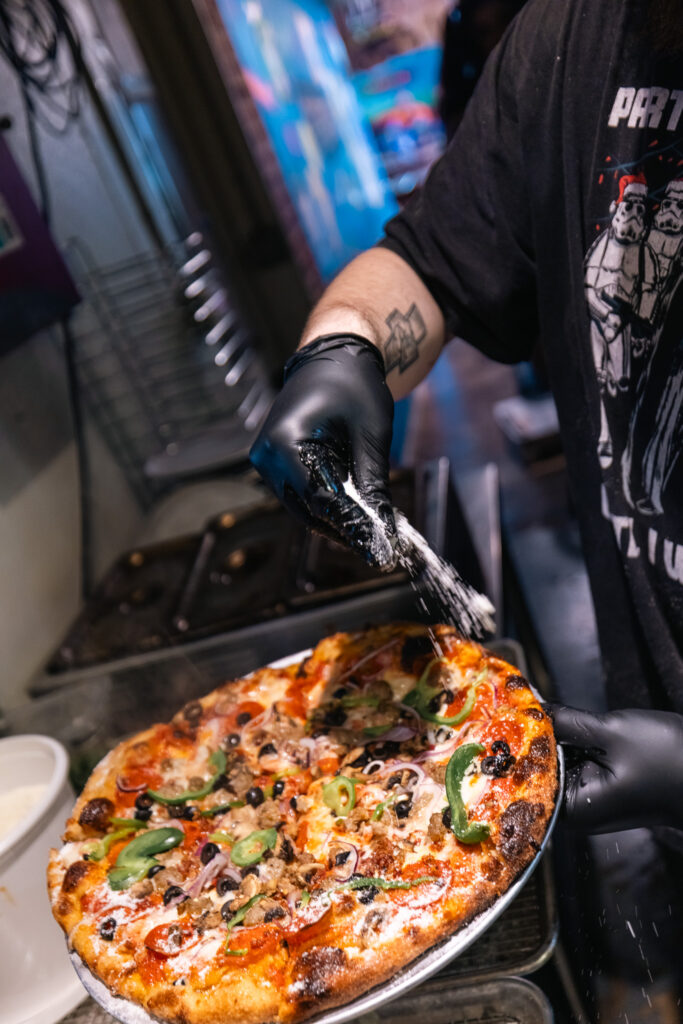 Employee at The Rail - A Pizza Company adds cheese to finished pizza.