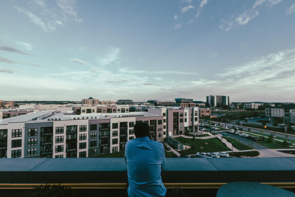 A person in a blue shirt looks out over an urban area with apartment complexes and parking lots from a rooftop balcony at dusk.