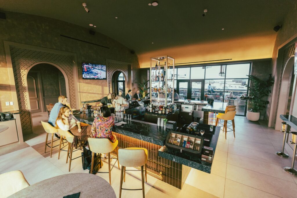 Patrons sit at a marble-topped bar in a lounge, with a television playing sports on the wall and a cigar humidor on the counter.