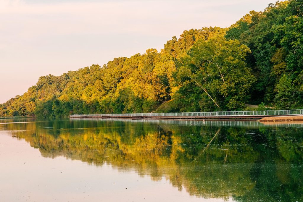 A calm river reflects green trees and a long fence-lined path under a clear sky, with sunlight illuminating the foliage.