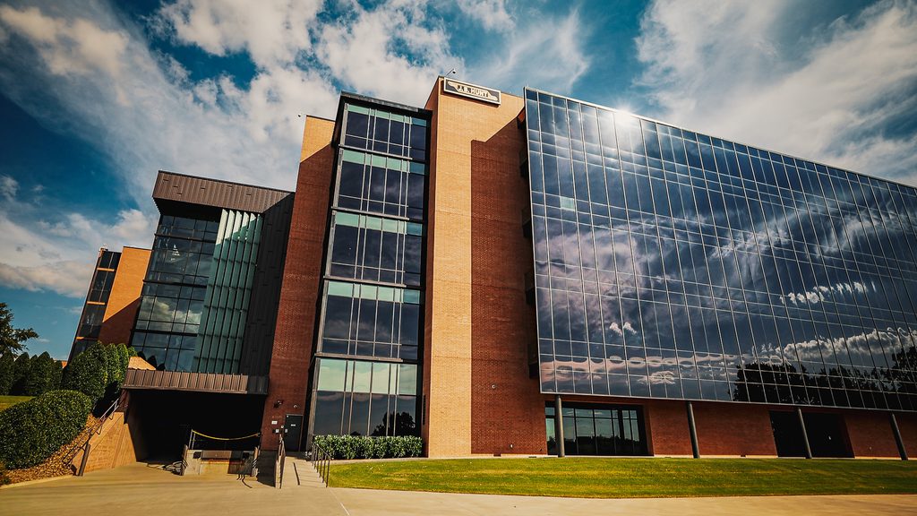 A modern glass and brick office building is shown under a blue sky with clouds, with reflective windows and a concrete walkway in front.