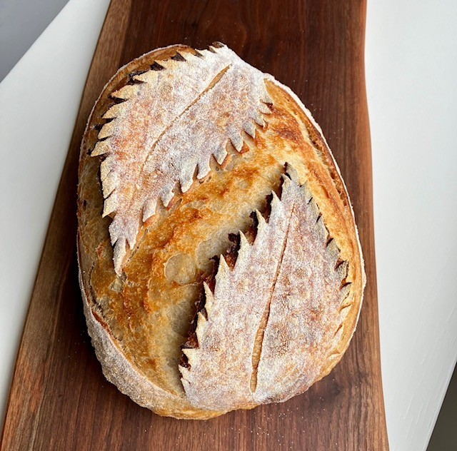 A round loaf of artisan bread with two large, leaf-shaped patterns scored on top, resting on a wooden board.