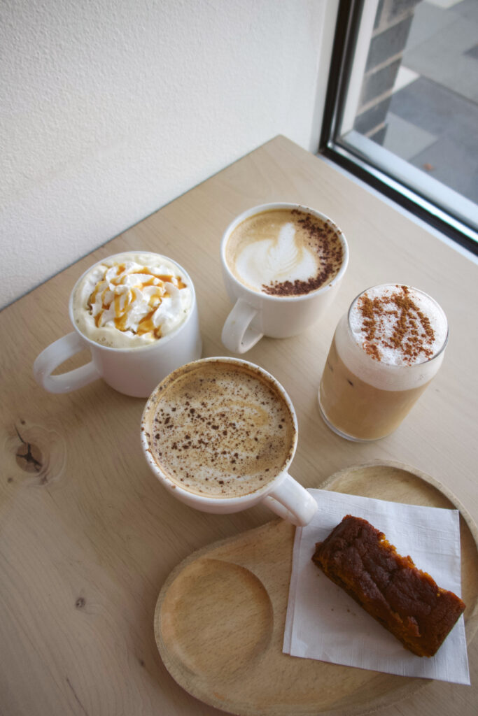 Four coffee drinks, two hot and two iced, are arranged on a wooden table next to a plate with a rectangular slice of cake, near a window.