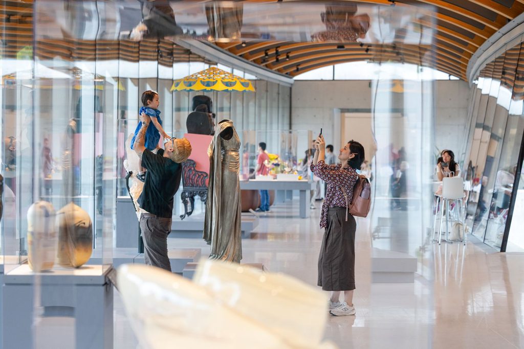 People inside a modern art museum; one person lifts a child, while another takes a photo. Sculptures and artifacts are displayed throughout the bright, glass-walled gallery.