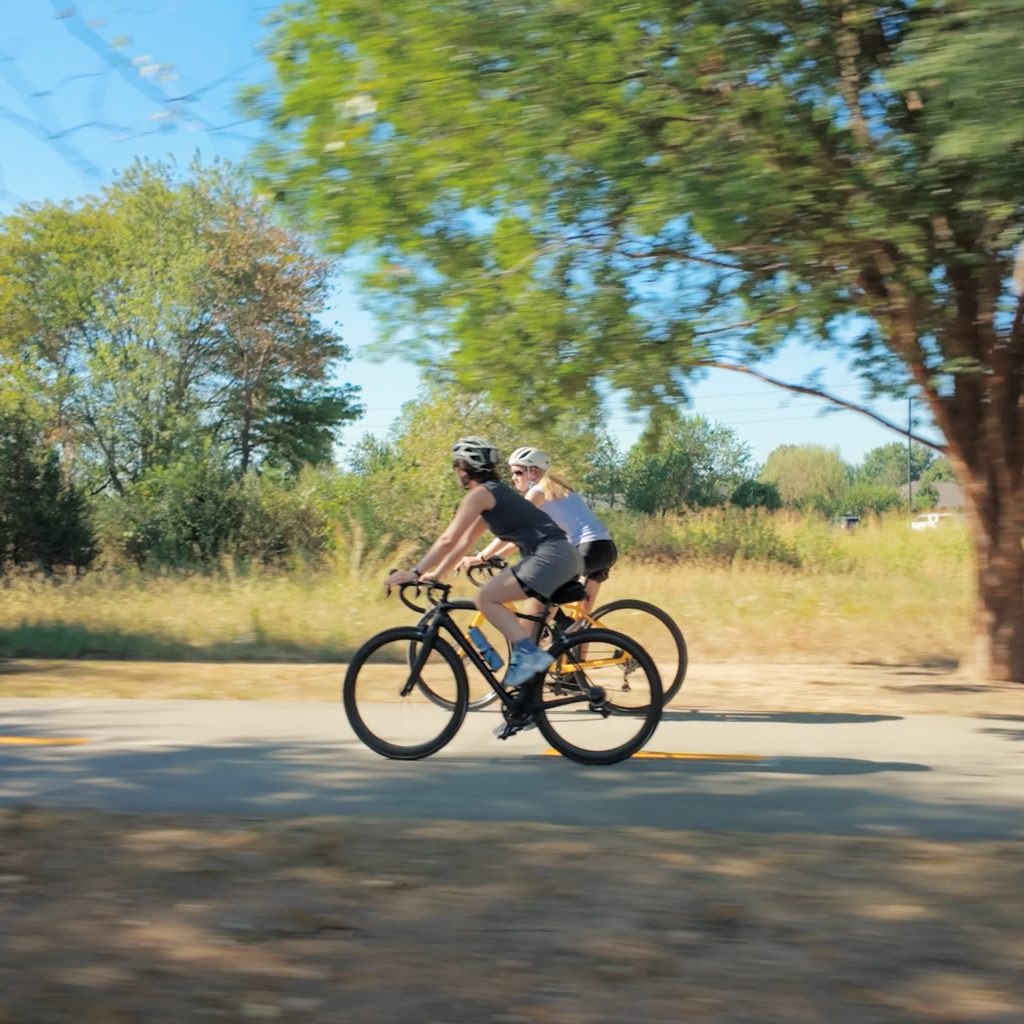 Two cyclists wearing helmets ride side by side on a paved path through a sunlit, tree-lined area.