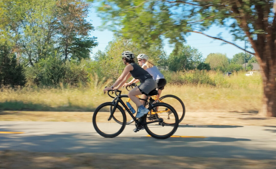 Two cyclists wearing helmets ride side by side on a paved path through a sunlit, tree-lined area.