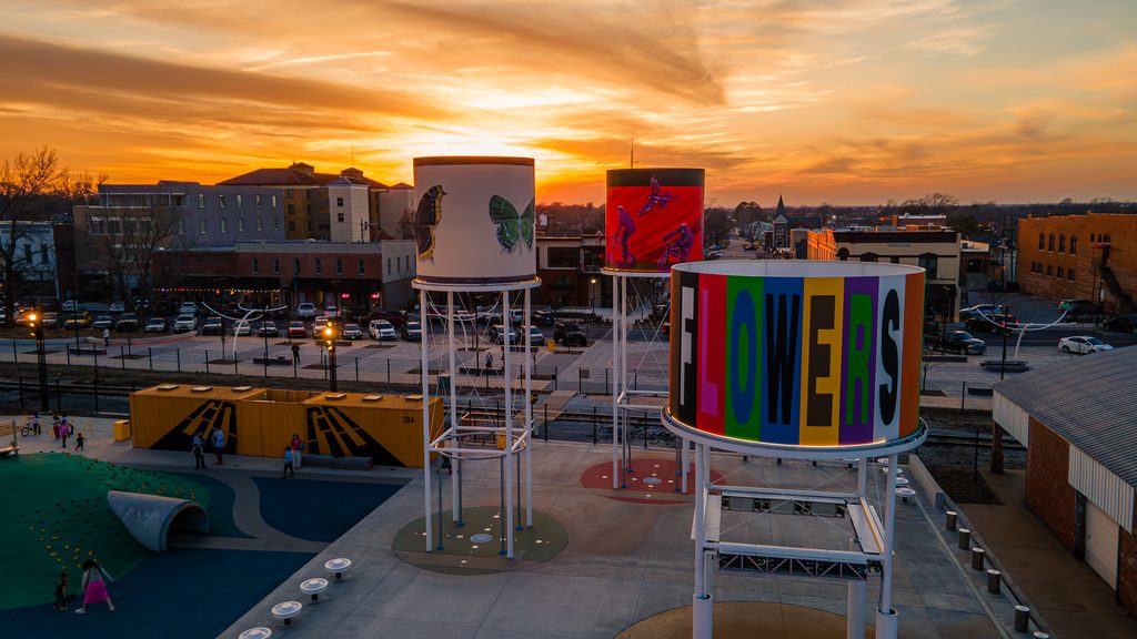 Three tall, colorful water towers with art and the word “FLOWERS” stand in an urban park at sunset, with buildings and a playground in the background.