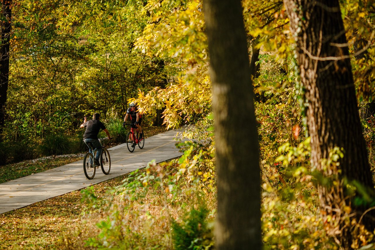Two people ride bicycles on a paved path on Razorback Greenway through a wooded area with autumn foliage.