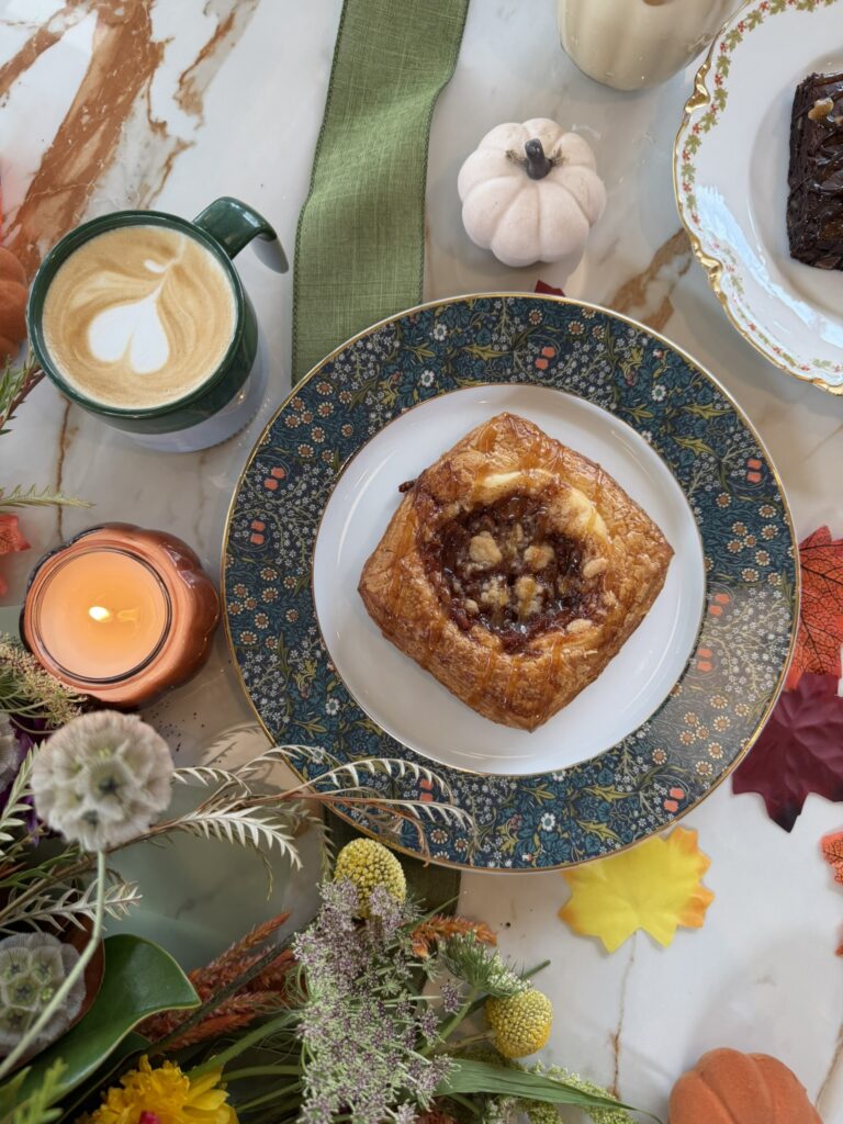A pastry on a decorative plate sits atop a marble table with a latte, candle, flowers, ceramic pumpkin, brownie, and autumn leaves arranged around it.
