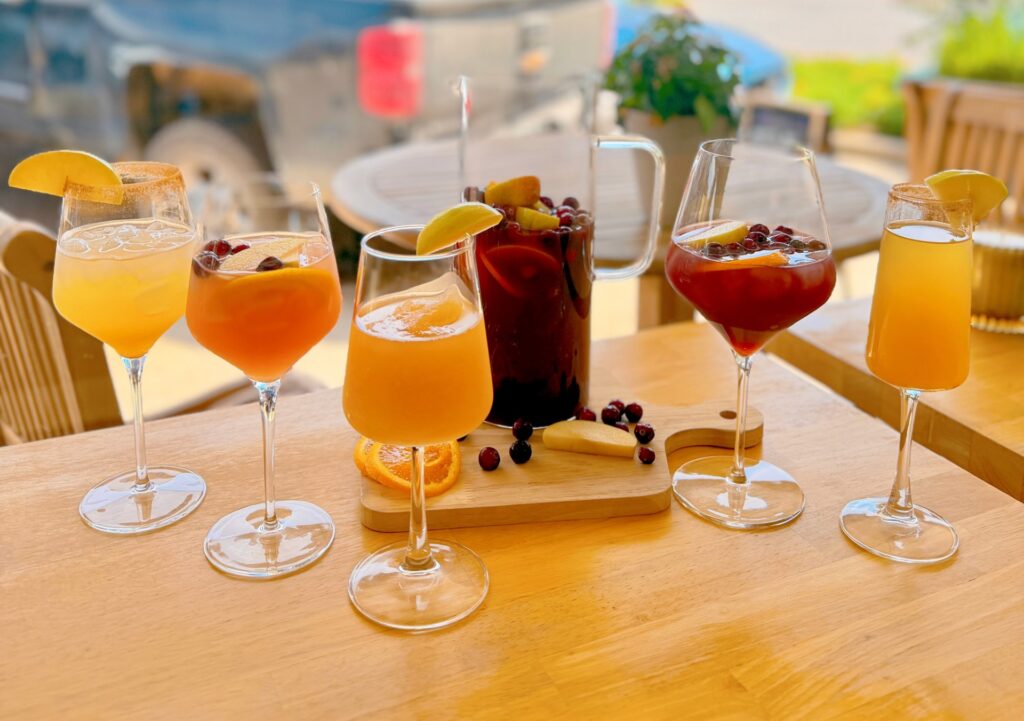 Six glasses of assorted fruit cocktails with garnishes are arranged on a wooden table, with a pitcher of red sangria and sliced fruit in the background.