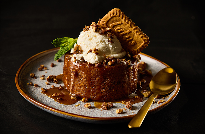 Sticky toffee pudding topped with a scoop of vanilla ice cream, a Lotus Biscoff cookie, and mint on a plate with a gold spoon.